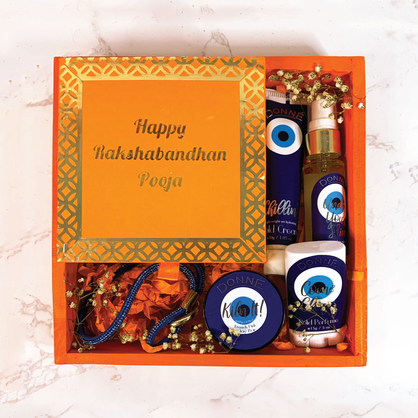 Assortment of products in a vibrant orange festive hamper with mithai scented candles, dazzling phone slings, moisturising body spray, portable solid perfume, lightweight facial moisturising cold cream and a french pink clay facial bar